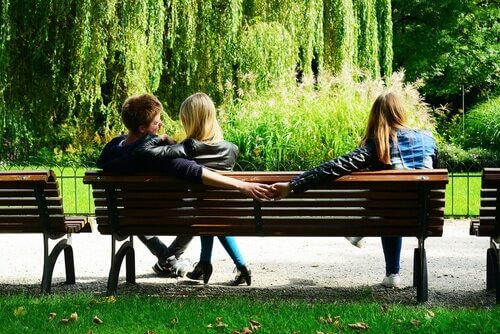 Uomo con compagna su panchina che si tiene per mano con altra donna di nascosto
